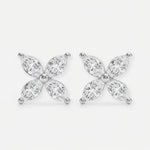 These two stud earrings are crafted from polished silver metal and set with clear, marquise-cut stones arranged in a four-petal floral shape. They are gently held between the thumb and index finger of a hand, showcasing their small, delicate size. The photo is shot against a softly blurred gray background with even natural light that highlights the sparkle of the stones. The overall style feels clean and minimal, focusing attention on the refined, symmetrical design.