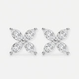 These two stud earrings are crafted from polished silver metal and set with clear, marquise-cut stones arranged in a four-petal floral shape. They are gently held between the thumb and index finger of a hand, showcasing their small, delicate size. The photo is shot against a softly blurred gray background with even natural light that highlights the sparkle of the stones. The overall style feels clean and minimal, focusing attention on the refined, symmetrical design.