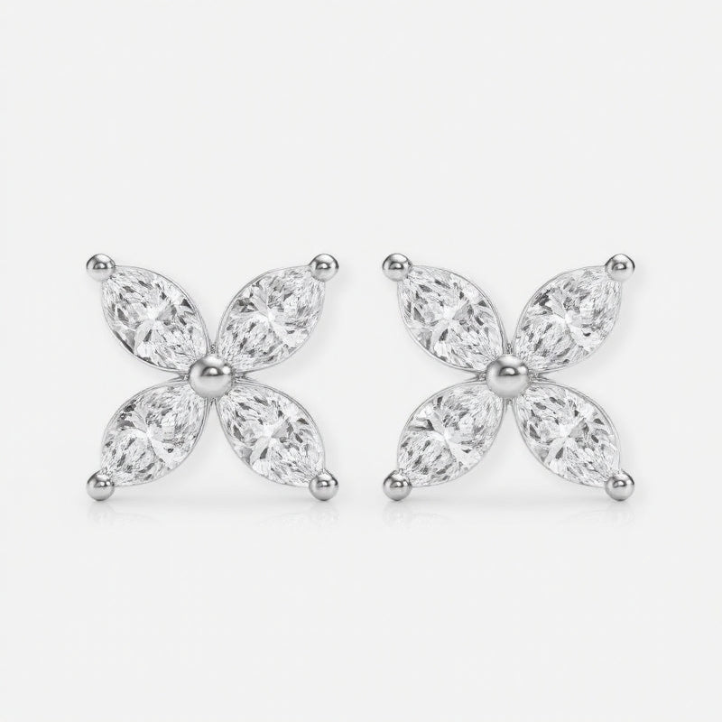 These two stud earrings are crafted from polished silver metal and set with clear, marquise-cut stones arranged in a four-petal floral shape. They are gently held between the thumb and index finger of a hand, showcasing their small, delicate size. The photo is shot against a softly blurred gray background with even natural light that highlights the sparkle of the stones. The overall style feels clean and minimal, focusing attention on the refined, symmetrical design.