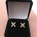 Two stud earrings made of silver-tone metal set with four clear marquise-cut stones form a delicate four-petal floral motif. They sit side by side on the black velvet cushion of an open black jewelry box, which rests on a soft pink surface. A gentle, diffused light highlights the facets of each stone against the dark interior, casting minimal shadows. The image has a clean, softly lit product-shot style with a slightly warm, inviting tone. | Adorn By You