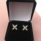 Two stud earrings made of silver-tone metal set with four clear marquise-cut stones form a delicate four-petal floral motif. They sit side by side on the black velvet cushion of an open black jewelry box, which rests on a soft pink surface. A gentle, diffused light highlights the facets of each stone against the dark interior, casting minimal shadows. The image has a clean, softly lit product-shot style with a slightly warm, inviting tone. | Adorn By You