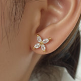A single gold stud earring featuring four marquise-cut clear gemstones arranged in a cross shape is set in yellow gold. It is worn on the model left earlobe and displayed in a tight profile headshot with dark hair pulled back. Soft, even lighting accentuates the sparkle of the stones against the wearer on smooth skin and white collar. The neutral background and crisp focus give the image a minimalist, editorial feel. | Adorn by You