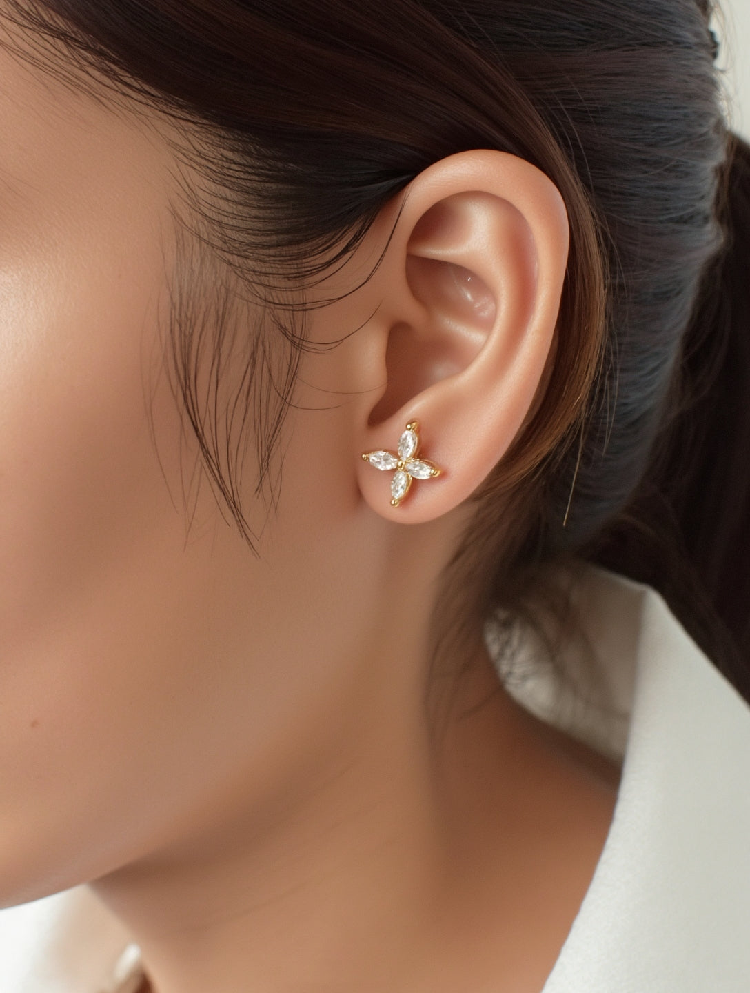 A single gold stud earring with four marquise-cut clear stones arranged in a cross-like floral pattern is shown snugly on the wearer\u2019s earlobe. Each stone is held in a delicate prong setting of warm yellow metal that gleams softly under diffused, natural lighting. The close-up shot captures fine details against a neutral backdrop of skin tones and dark hair, giving the image a minimalist, editorial feel.| Adorn By You