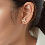 A single gold stud earring with four marquise-cut clear stones arranged in a cross-like floral pattern is shown snugly on the wearer\u2019s earlobe. Each stone is held in a delicate prong setting of warm yellow metal that gleams softly under diffused, natural lighting. The close-up shot captures fine details against a neutral backdrop of skin tones and dark hair, giving the image a minimalist, editorial feel.| Adorn By You