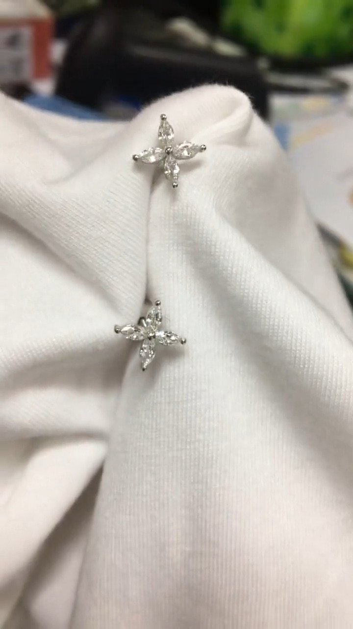 Two silver stud earrings with four clear, marquise-cut stones set in a prong-style cross are nestled into a crisp white fabric. The gemstones sparkle softly under diffused, natural light, their facets catching gentle highlights. Captured in a close-up with a shallow depth of field, the earrings are displayed pinned to the folds of the cloth against a softly blurred indoor background. | Adorn By You
